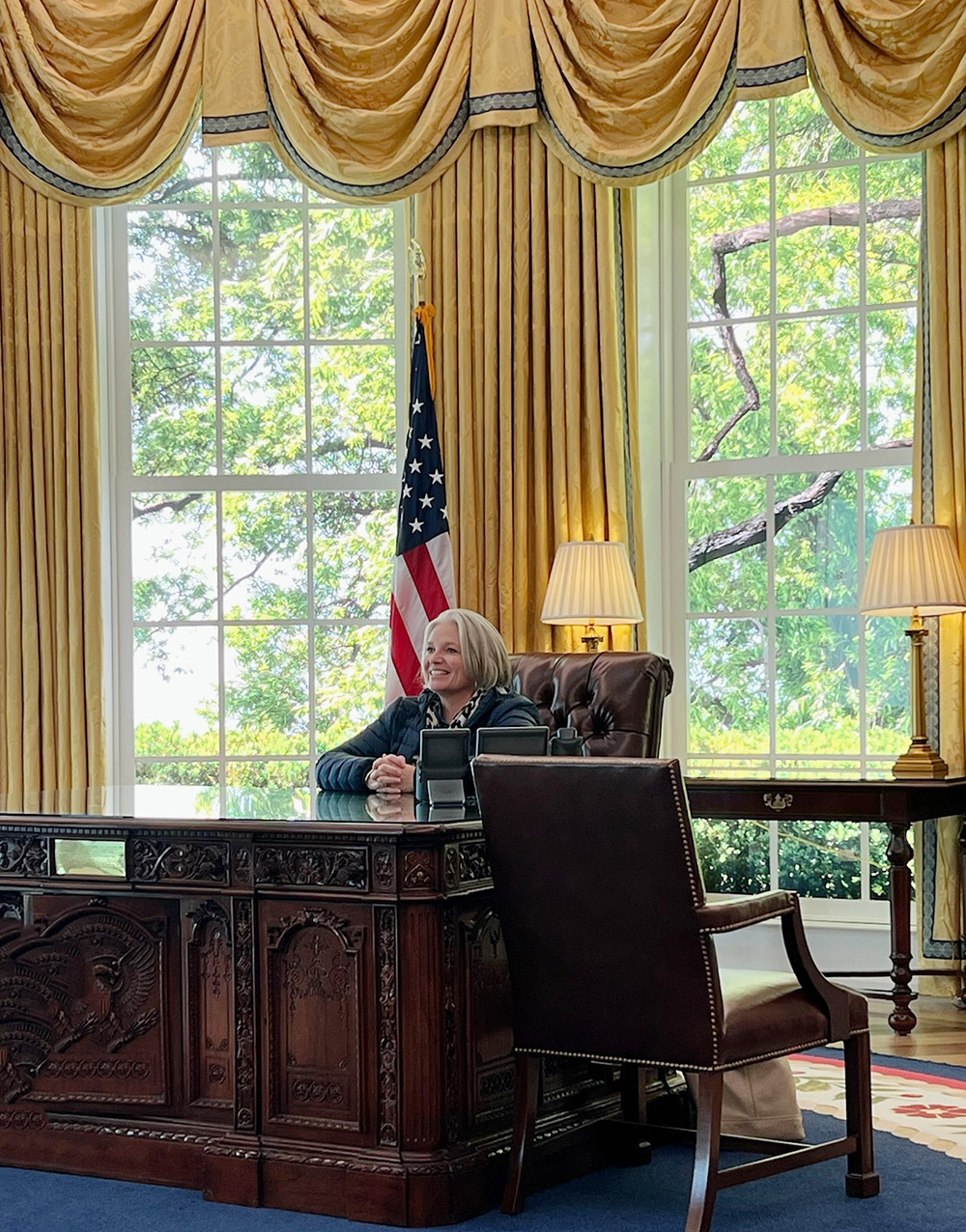 Megan smiles while sitting at the desk in the Oval Office.